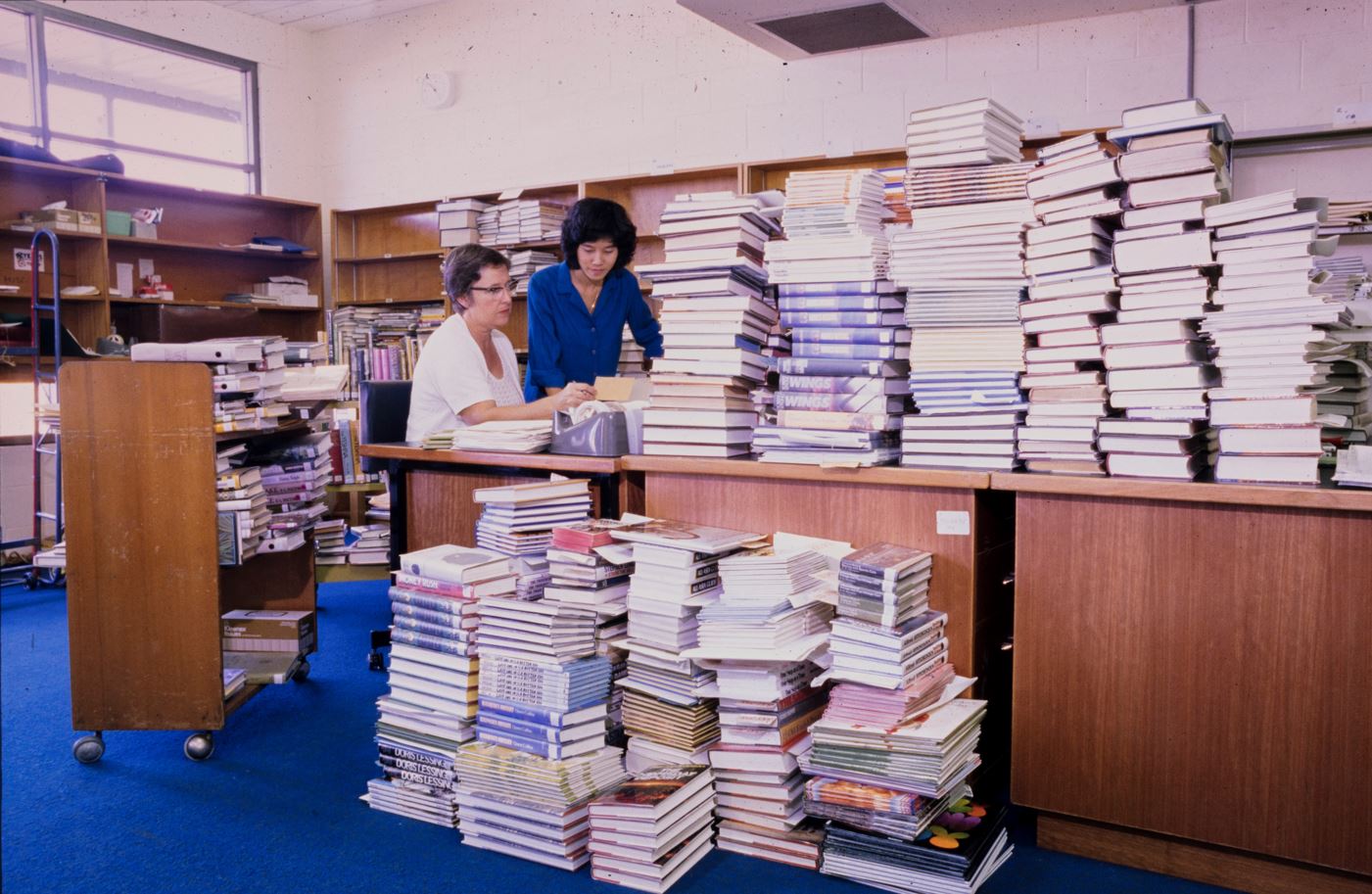 Two staff members at a desk covered in books 1982