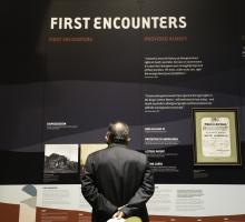 A man viewing an exhibition. A label on the wall reads First encounters