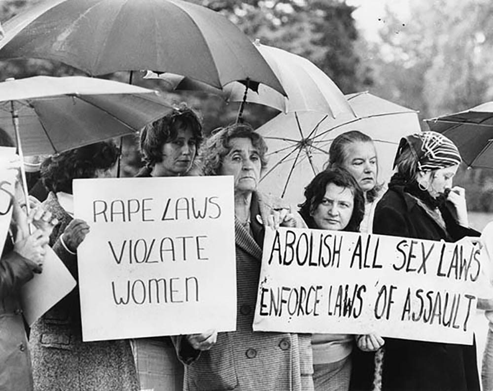 Group of women protesting in the rain, holding umbrellas and signs that say: 'Rape laws violate women' and 'abolish all sex laws enforce laws of assault'