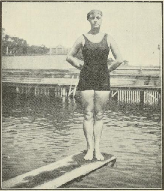 Black and white image of a woman in swimwear standing on a diving board in 1916.
