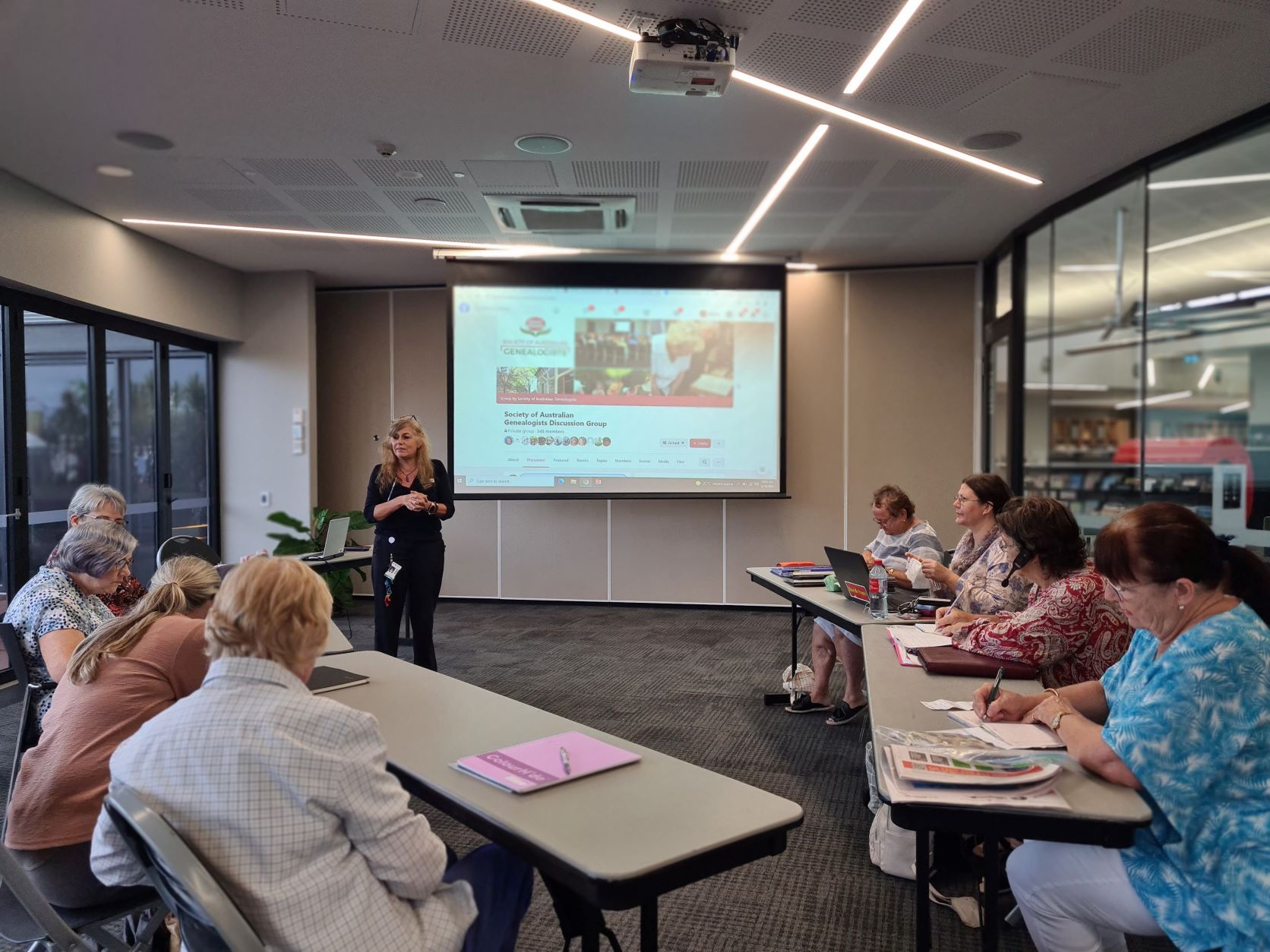 A woman presents in front of a projector screen to a group of women learning