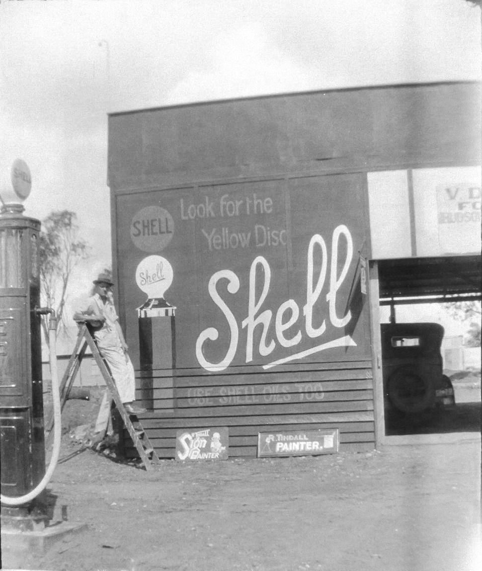 A black and white image at a petrol station in the 1940's. There is a petrol bowser on the left of the screen and next to it is a man resting against a ladder. He is positioned in front of a large wall with a hand-painted advertisement for Shell Petroleum which reads 'Look for the yellow disc'. 