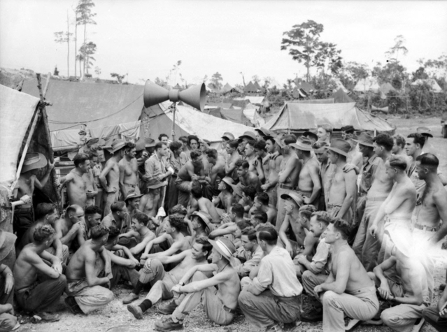 A large group of soldiers sit and stand in a semi circle. Amongst them stands Corporal R L Stokes as he presents the race. Behind them are a series of tents an amplifier is above them. Many of the soldiers are topless in the heat of New Guinea