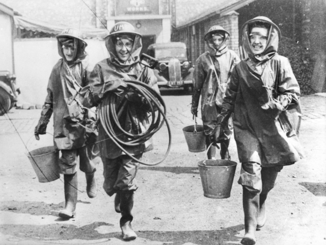 four women wearing full firefighting gear, including rubber boots and helmets, walking in unison towards the camera. They are holding hoses and buckets and are smiling broadly.