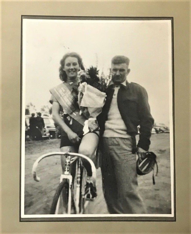 Iris Bent posing for a photograph on her bike holding a bouquet of flowers. There is a man posing with her.
