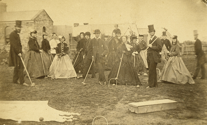 Group of men and woman posed with croquet sticks. The men are wearing suits and top hats and the women are wearing crinoline dresses