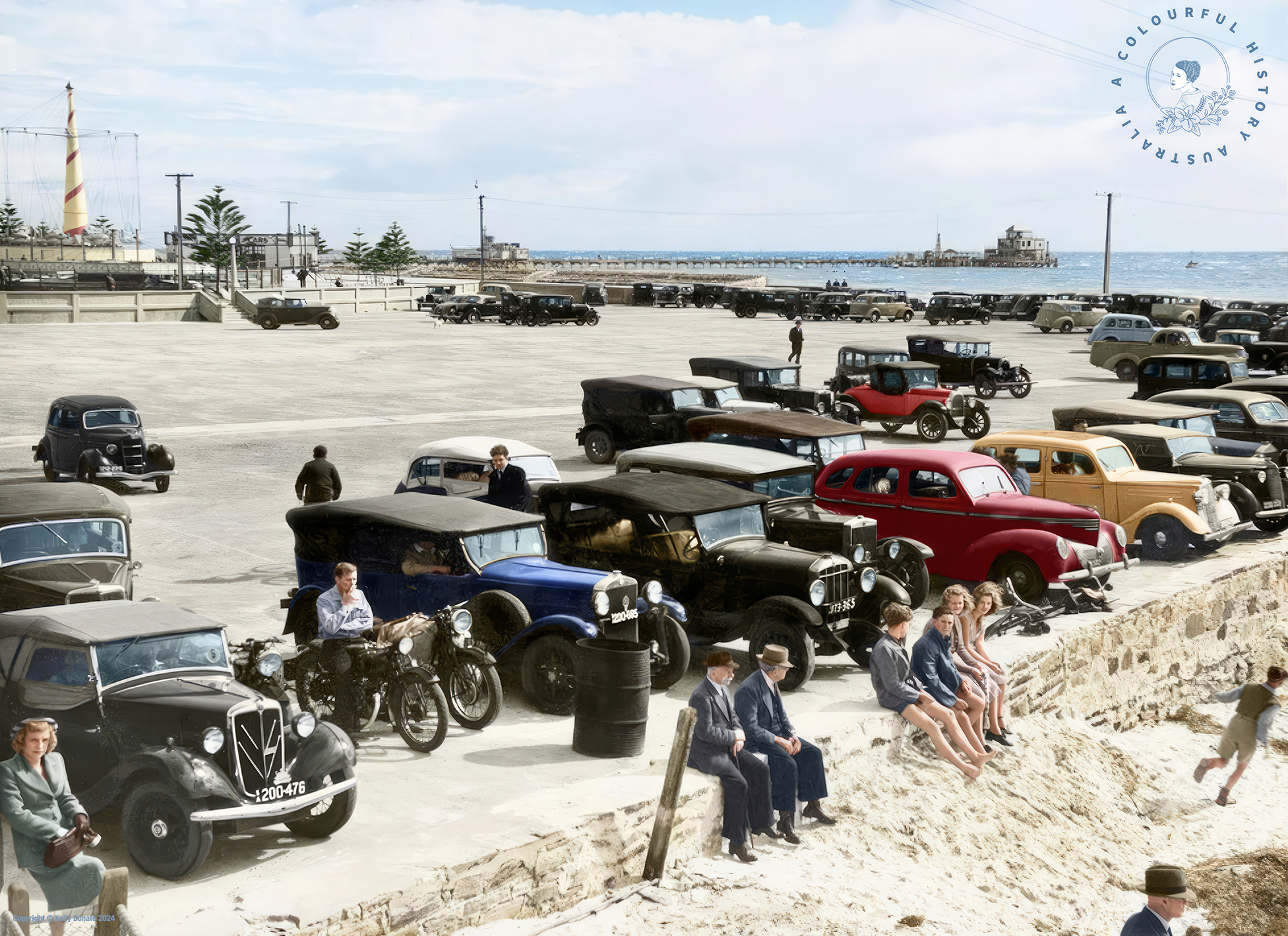 colourised image of historical cars parked at a beach. Beachgoers sit on a ledge next to the sand in front of the cars and the ocean can be seen in the background.