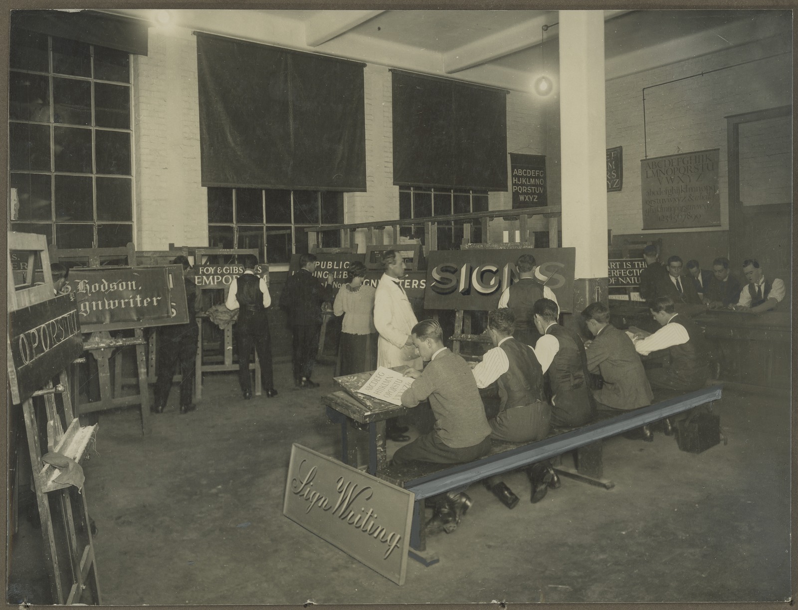 A sepia image of a signwriting class with fourteen male students, one female student and a male teacher. Five students are positioned on a large bench at the forefront of the image. Six students are positioned in front of easels, hand painting signs. The other students are to the back right of the image, sitting around a large table. 