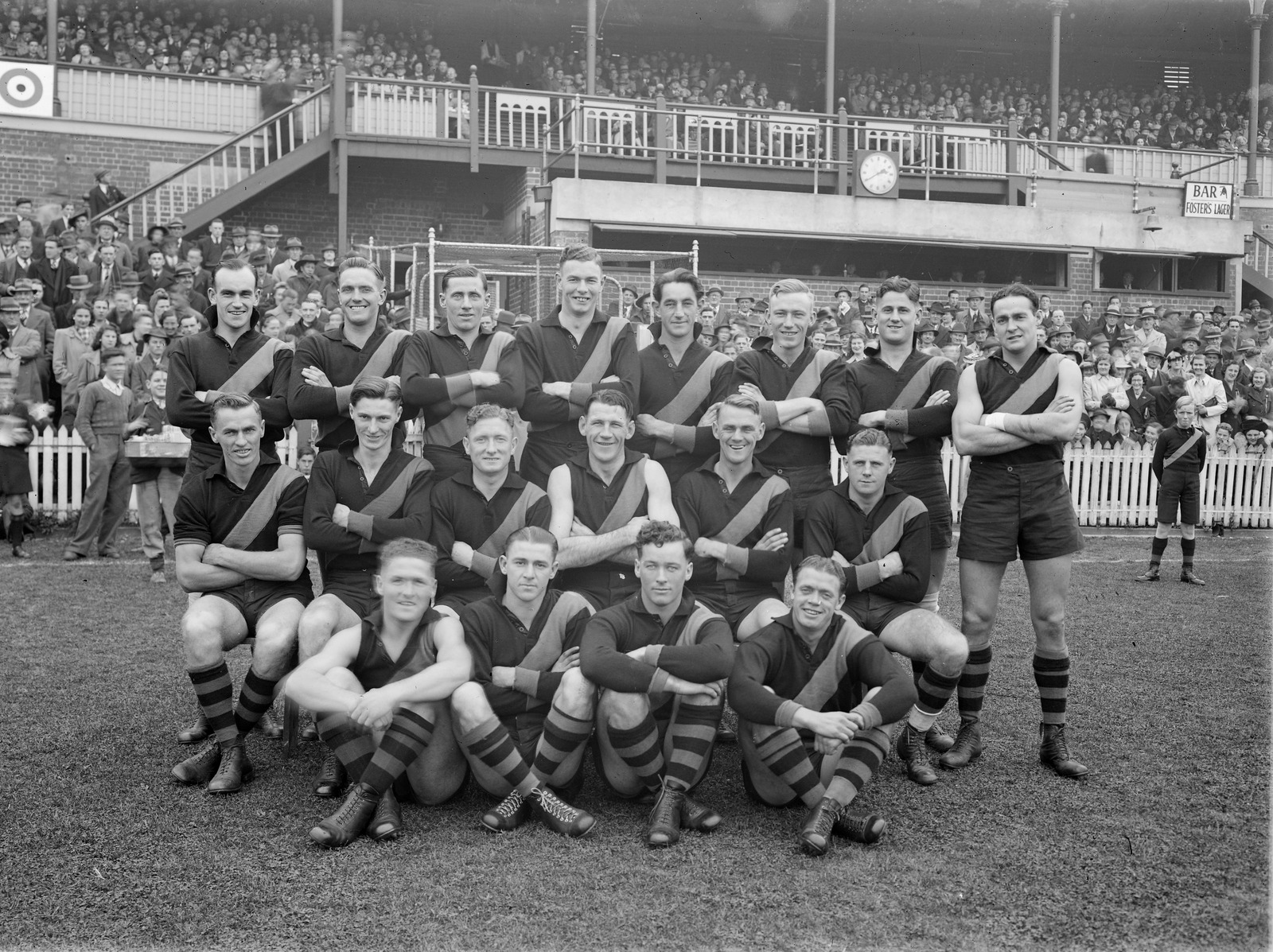 Richmond Football Team (18 players) pose for a photo in from of a grandstand full of fans.