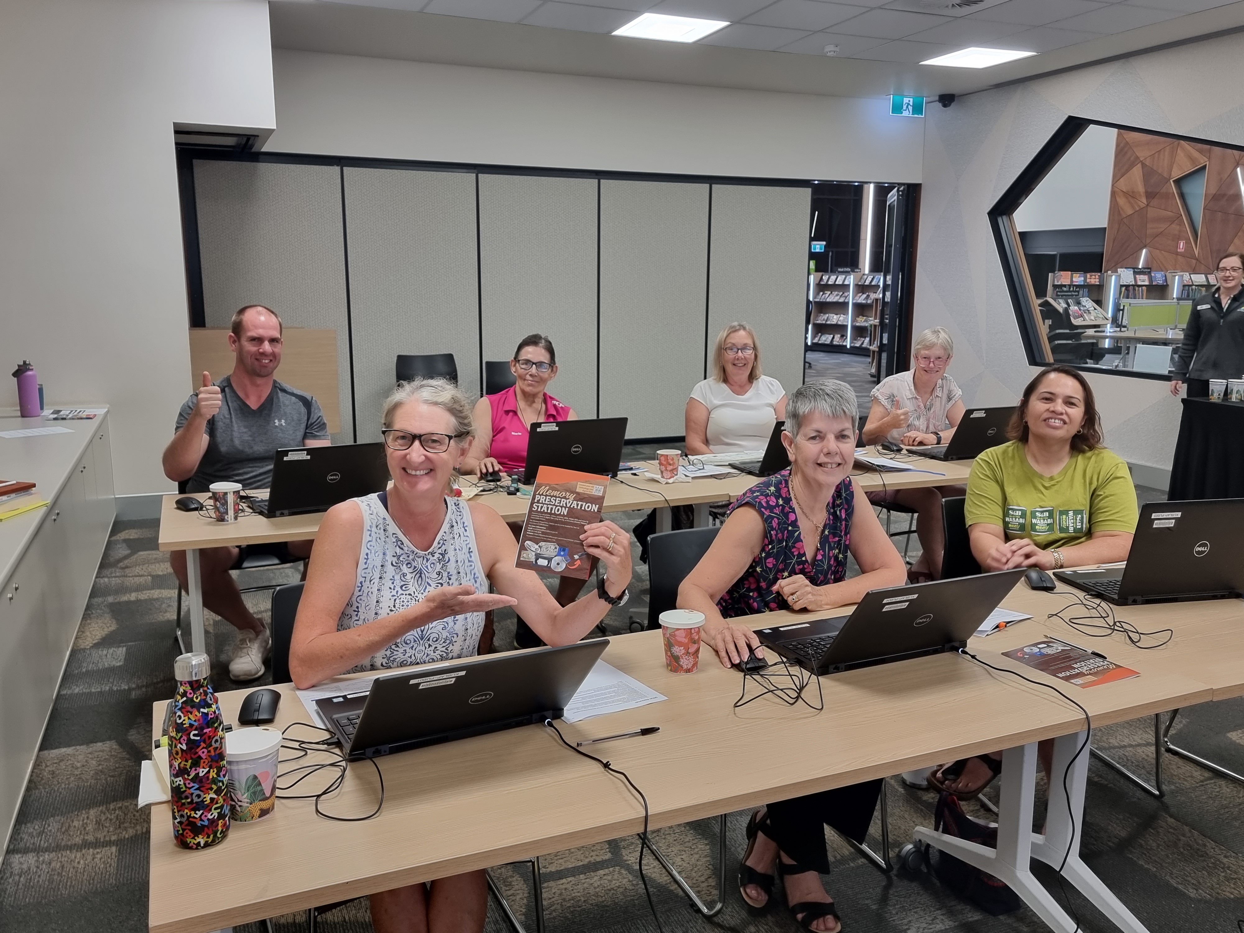 A group of people smiling, sitting at desks with computers and looking at the camera.