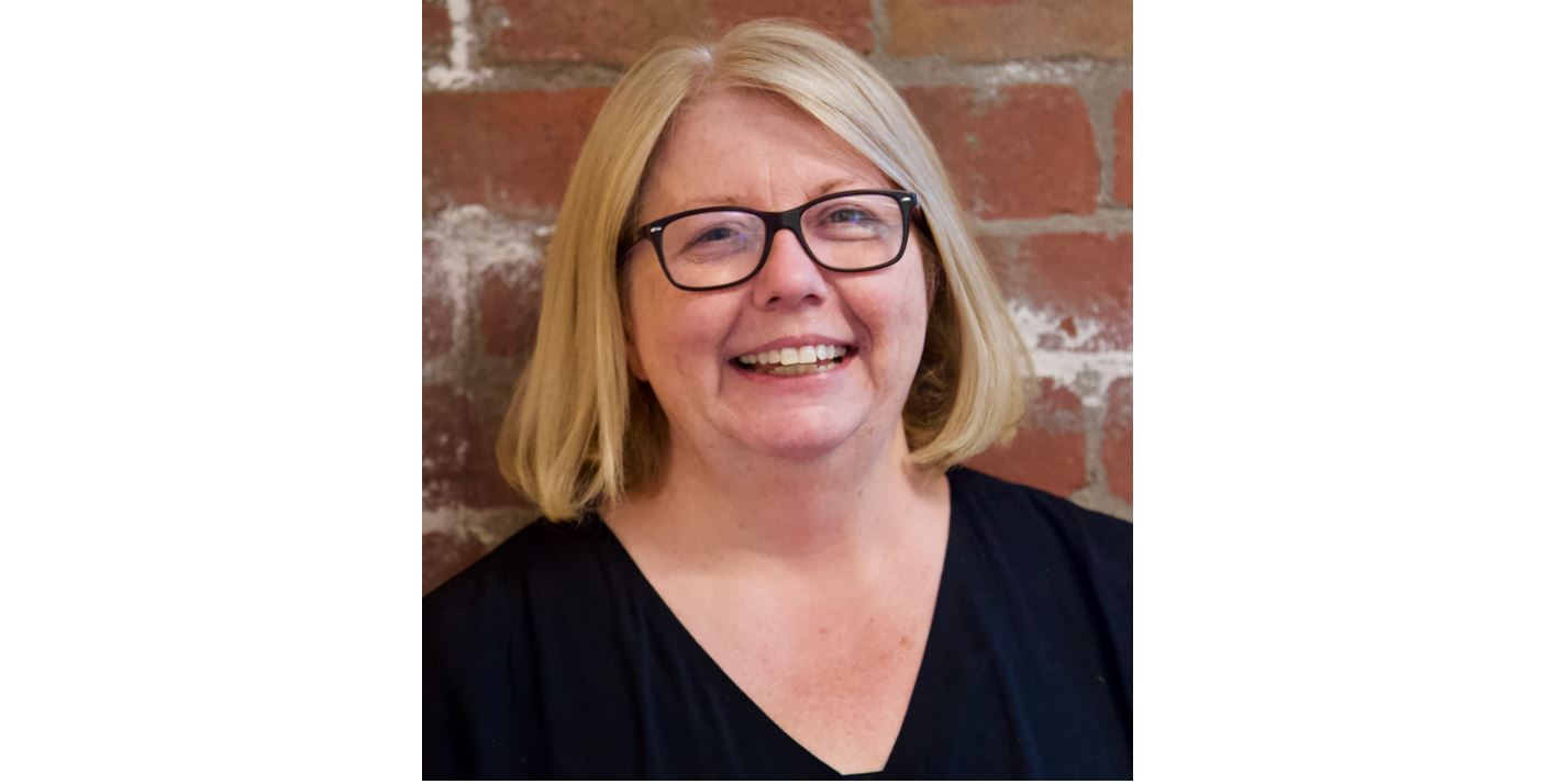 A headshot of a woman with blond hair, black framed glasses and a wide, welcoming smile.