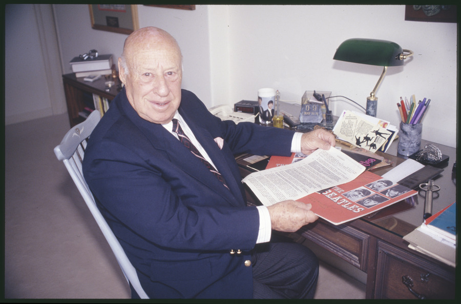 An older man wearing a blue suit and maroon tie sitting at a work desk holding the Beatles Australian Tour contract and other Beatles memorabilia.
