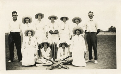 A club photo with women in the centre wearing hats and dresses, cricket bats at their feet, two men stand either side of the group.