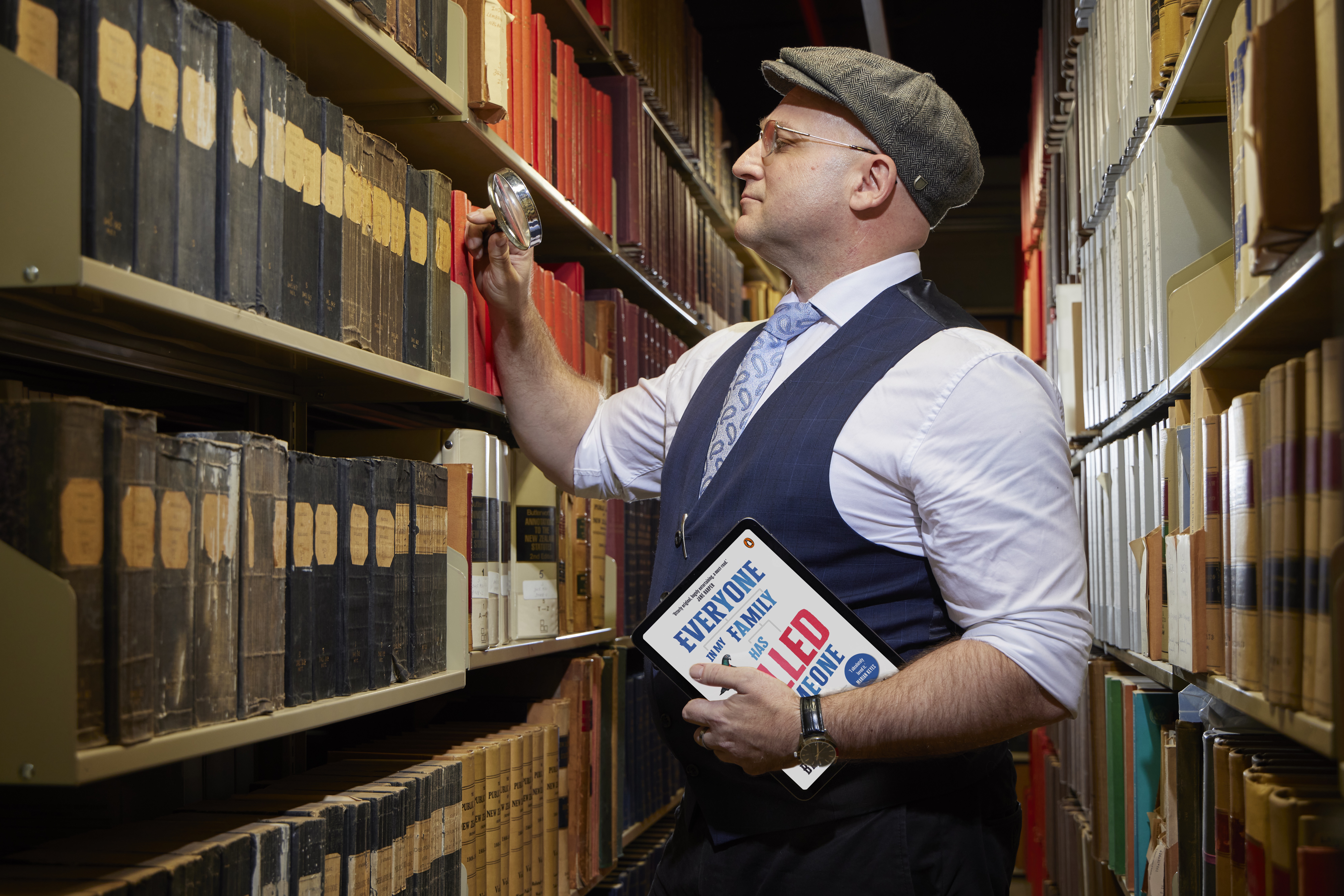 Man standing in the stacks/shelves of the Library wearing a button up shirt, tie, waistcoat, and newsboy cap holding a tablet displaying a book cover in his left hand and holding a magnifying glass up to the shelves. 