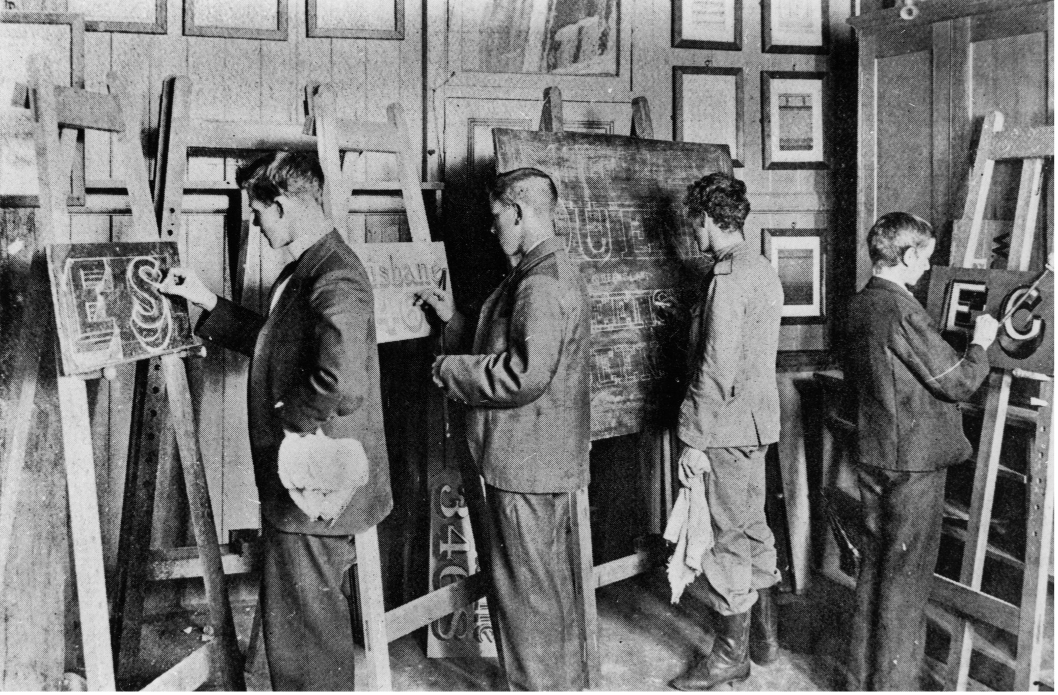 A black and white image of three men and one woman in a songwriting class. They all have an easel in front of them and they are all midway through painting a sign. 