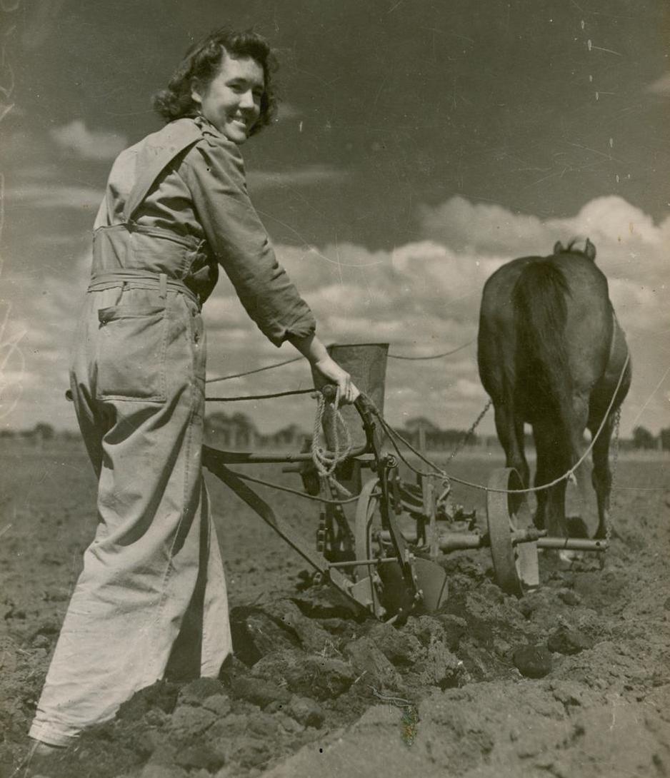 Women pushing a plow behind a horse in a muddy field. She looks back towards the camera smiling