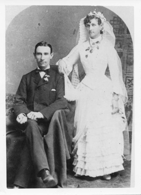 A man wearing a tuxedo and a woman in a wedding dress. The man is sitting and the woman standing behind him with her hand on his shoulder.