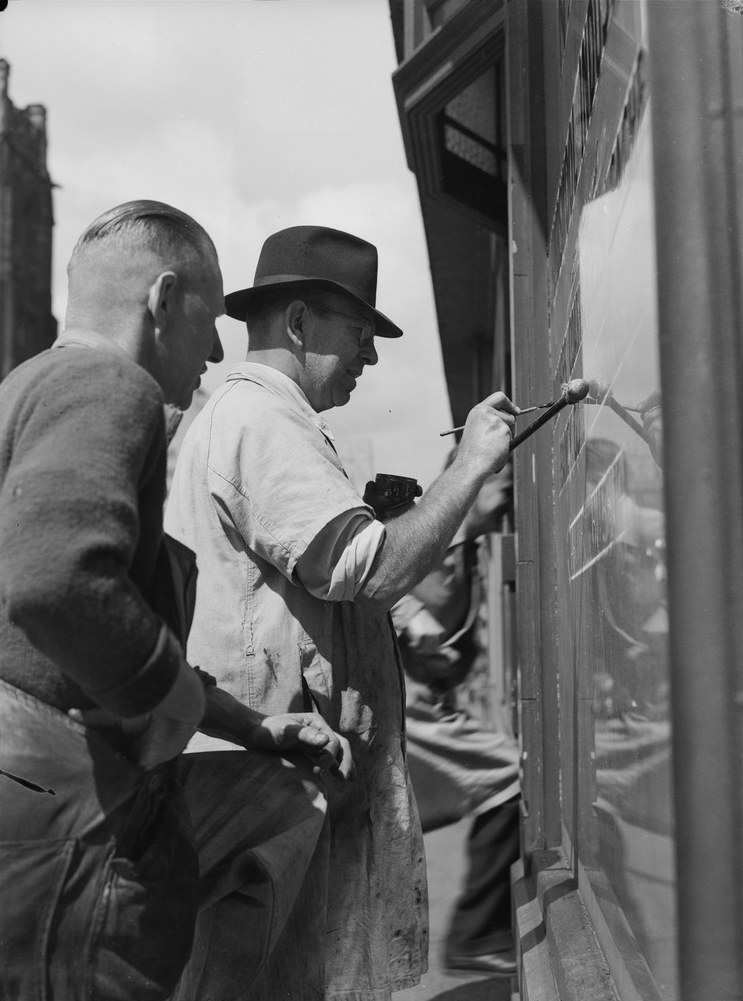A black and white image of two men. One of the men is painting on a window while the other man watch closely over his shoulder. 