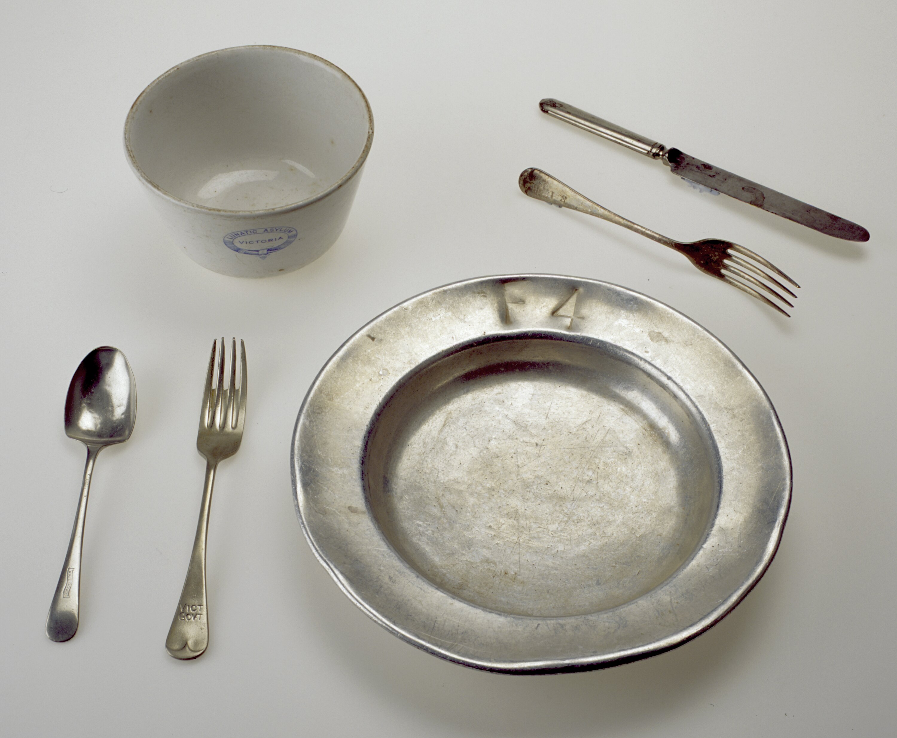 A metal plate and cutlery set. There is a ceramic cup to the side with the wording Beechworth Lunatic Asylum.