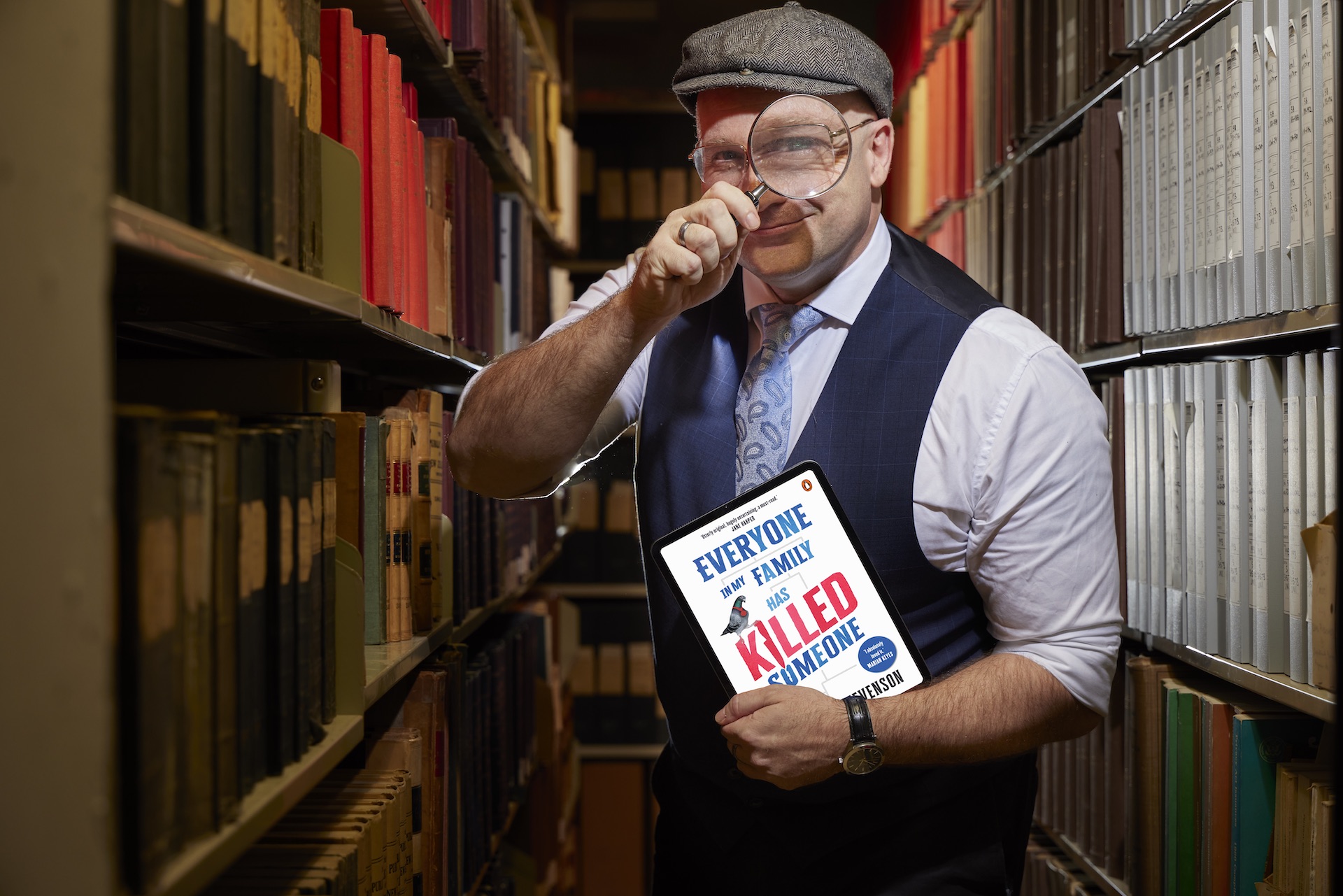Man standing in the stacks/shelves of the Library wearing a button up shirt, tie, waistcoat, and newsboy cap holding a tablet displaying a book cover in his left hand and holding a magnifying glass up to his eye with his right hand. 