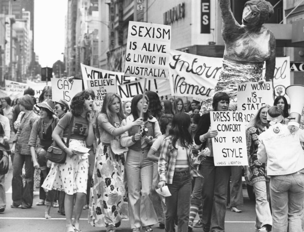 Large group of women walking down the middle of a road protesting with signs and a giant paper mache women sculpture 