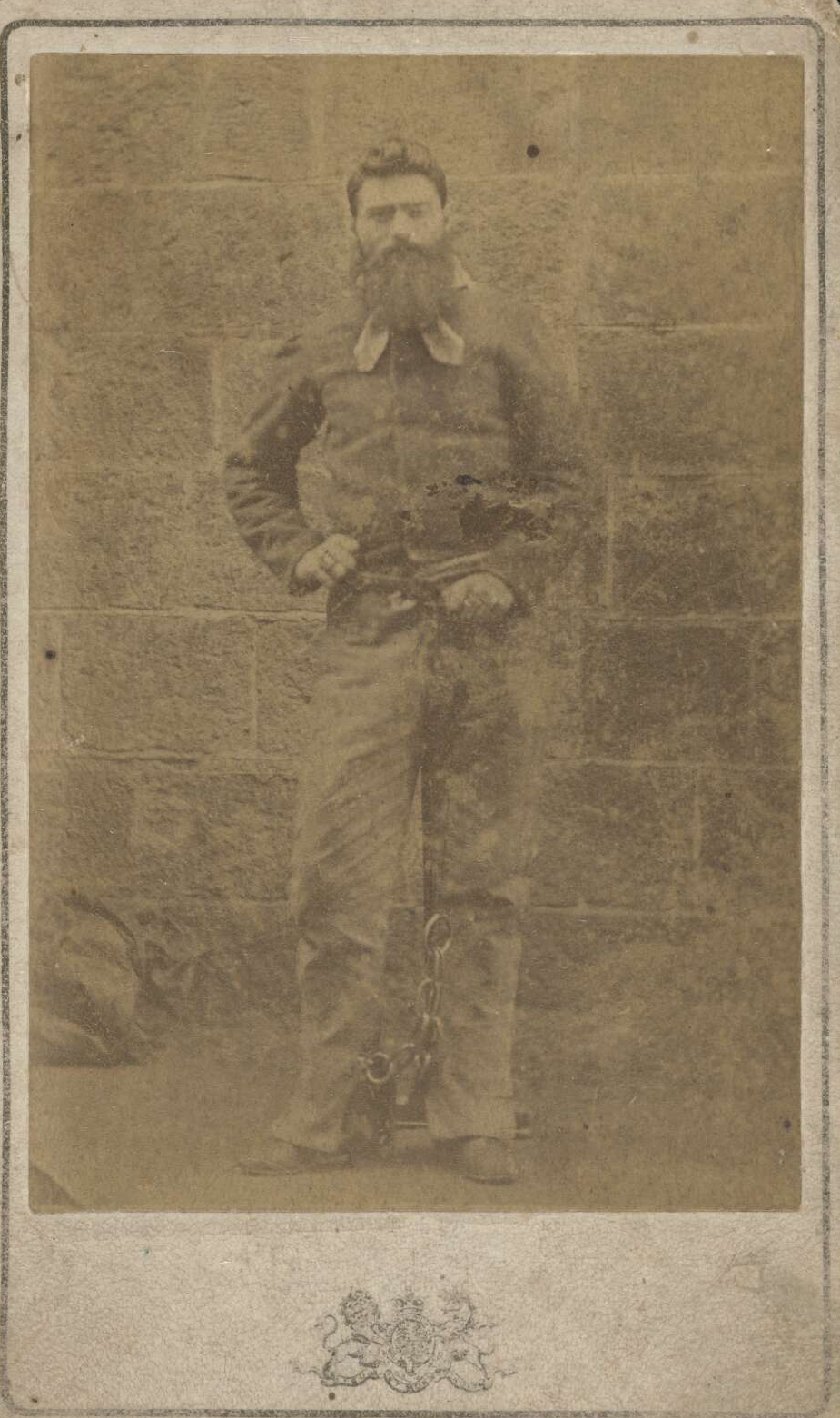 Ned Kelly standing in front of a concrete block wall.