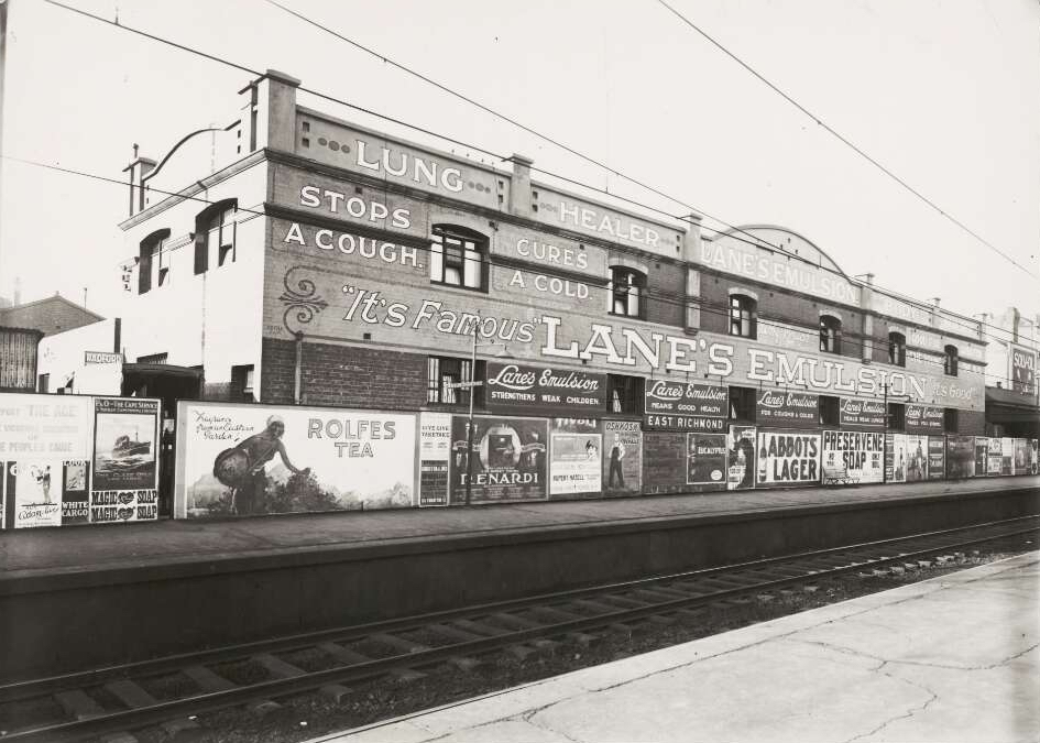 A black and white image of a large two-storey building next to train tracks. The building is covered in advertising signage with the largest sign for Lane's Emulsions, spanning across the whole building. 