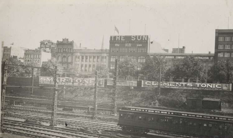 A sepia landscape image of Flinders Street in Melbourne in the 1930's. There are railway lines at the forefront of the image. Further back there is fencing in front of buildings with painted signage running along the length of the fence. The visible advertising is for Smartone for suede shoes and Clements Tonic. The Sun building is at the centre of the image. 