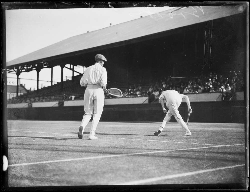 ir Norman Brookes playing a doubles tennis game. Behind him a crowd of spectators watch on from the stands.