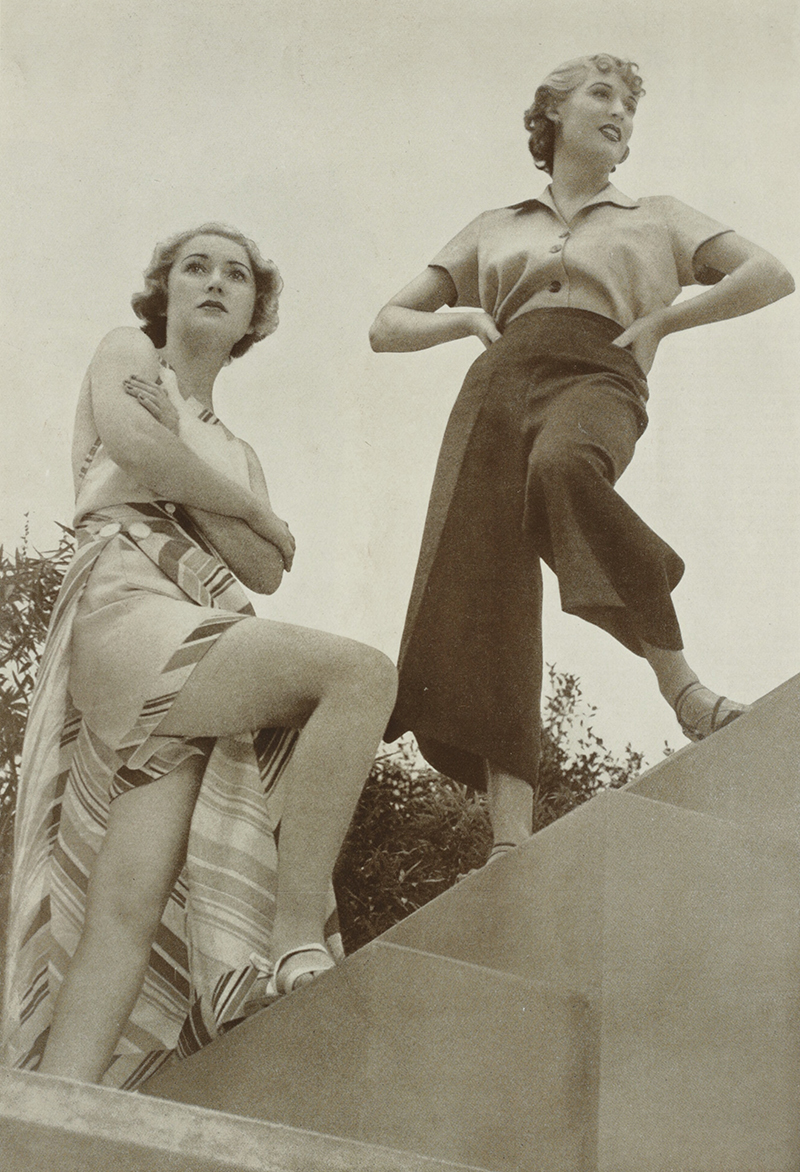 Two women standing on stairs. One is wearing loose pants and buttoned blouse. The other is wearing shorts with a wrap-around skirt on top.