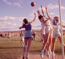 A coloured image of a netball game. Four girls in uniforms leap towards a ball around a goalpost.