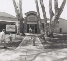 A community building with people coming in and out and some trees around the entrance. 