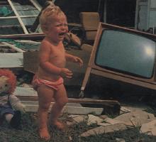 A child crying in the wreckage of a destroyed home with a tv and a toy in the background.