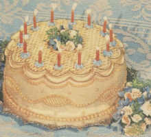 A yellow birthday cake with candles on a blue and white table cloth with a bunch of decorative flowers to the right. 
