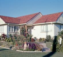 a small post war house with a red roof surrounded by lush garden beds